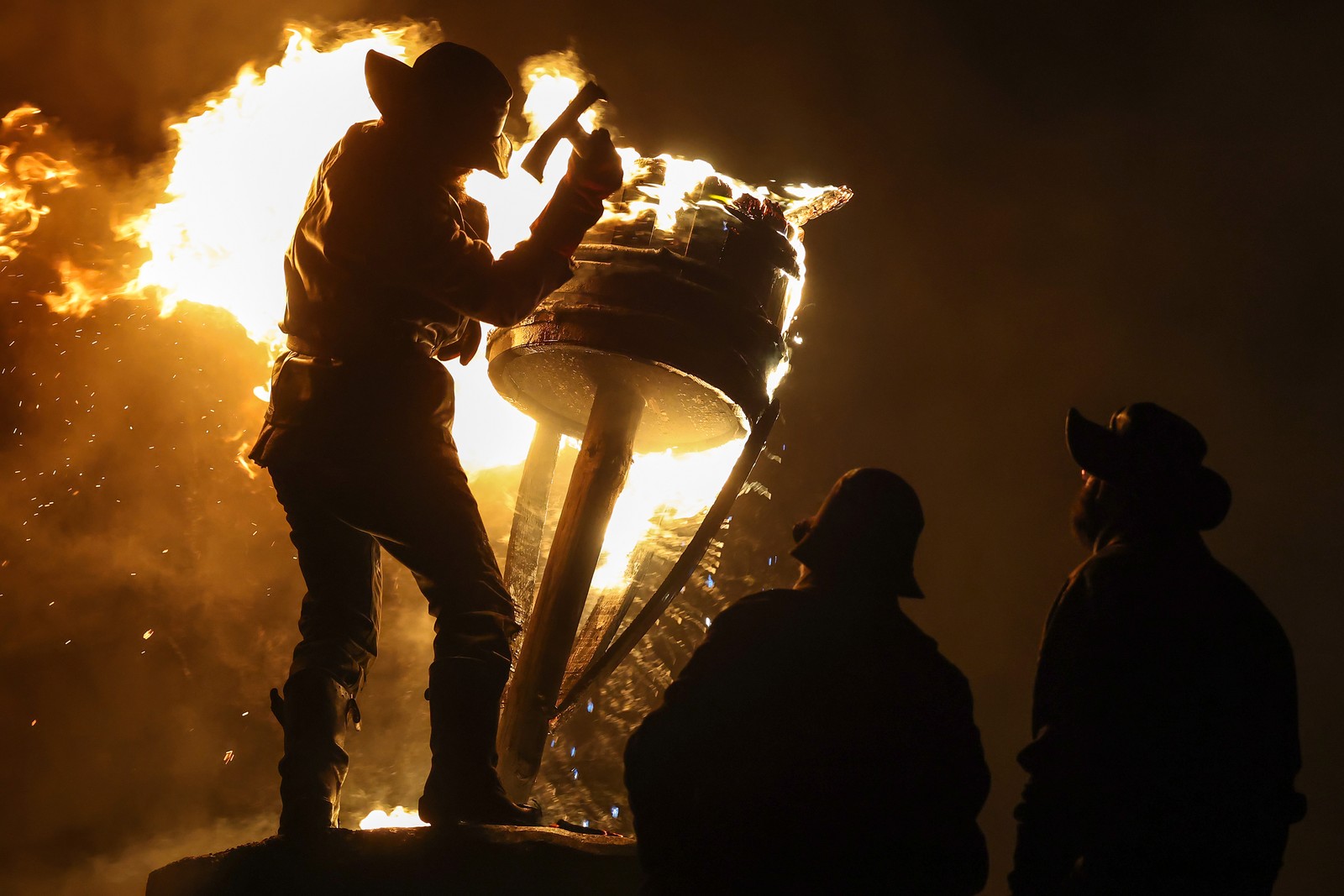 A person wearing protective gear holds a small axe beside a flaming barrel, during a festival.