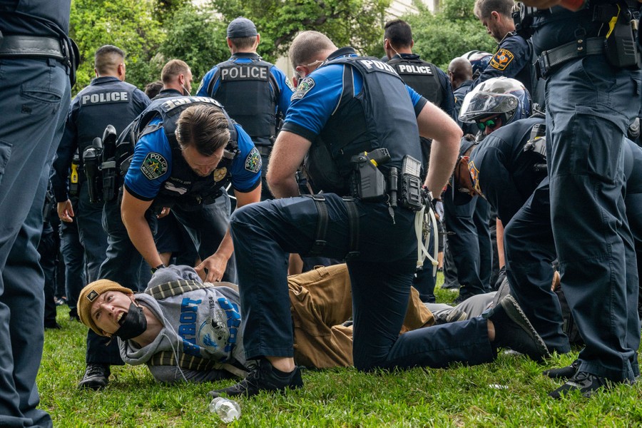 A large number of police officers stand around on grass while two officers kneel down to arrest a protester who is lying on the ground.