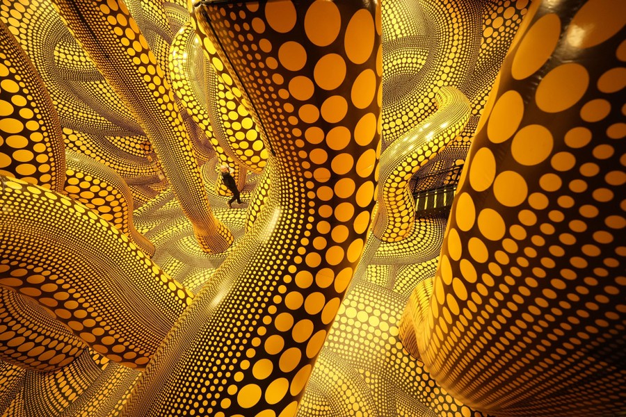 A person walks through an art installation—a large room filled with long, inflatable, tentacle-like tubes. Everything is painted with yellow dots on a black background.