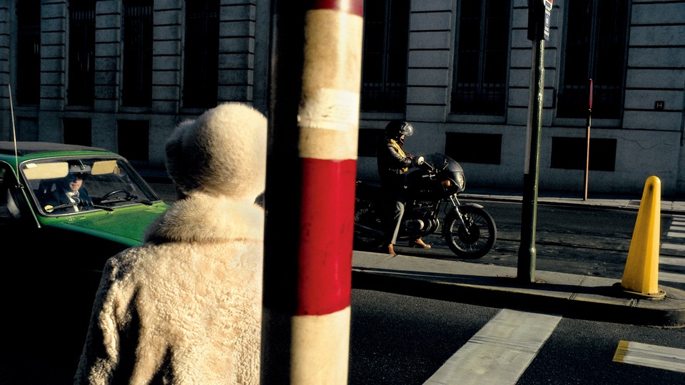 a person in a white fur coat and hat faces away from the camera, toward a green car on the street with a man inside