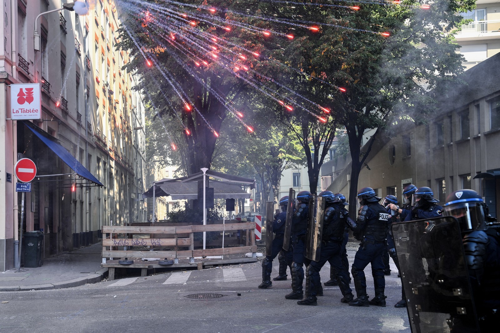 Riot police officers stand in a street as fireworks go off above them.