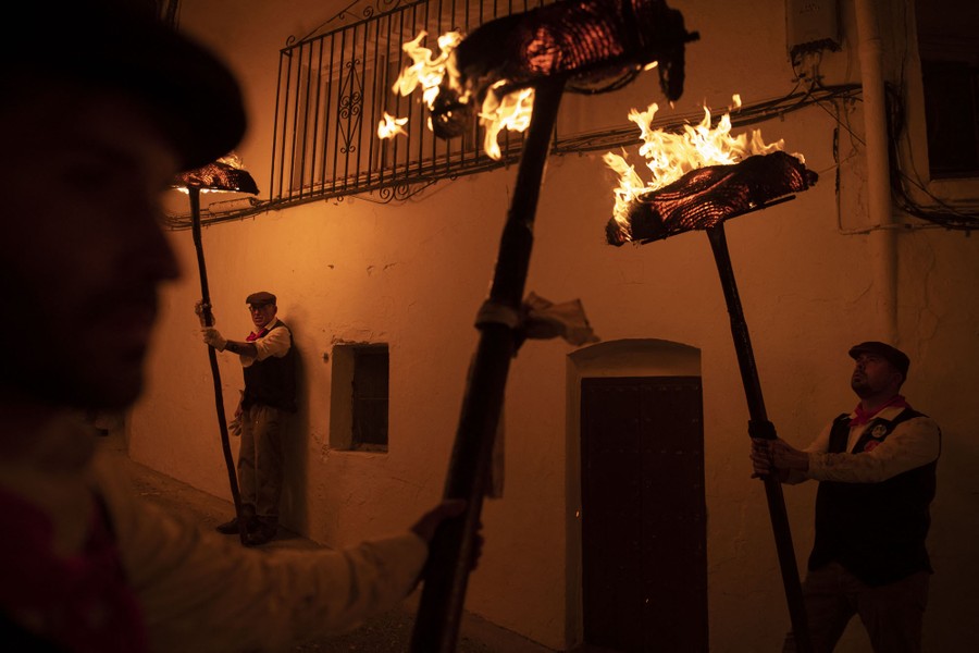 Several people in traditional clothing stand in a narrow street, holding long sticks with flaming bundles at the end.