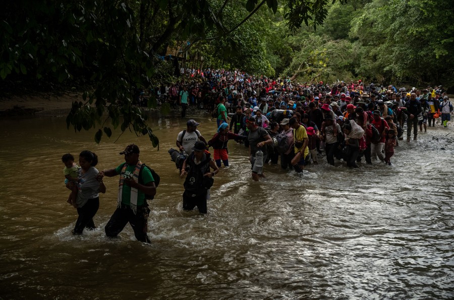 A large group of migrants stands along the edge of a jungle river, with many starting to wade across.