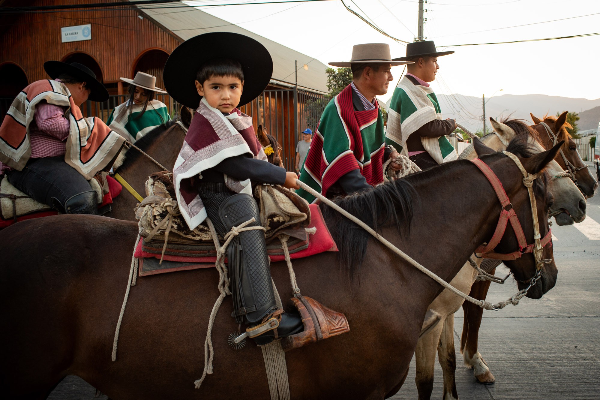 A boy and several adults ride horses while wearing traditional clothing.
