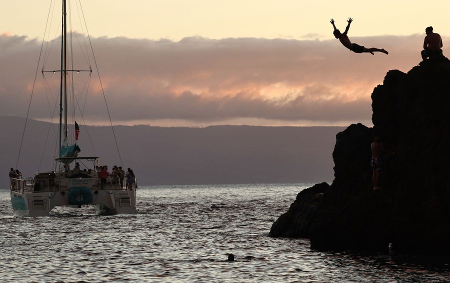A person dives off rocks as a tourist boat passes nearby.