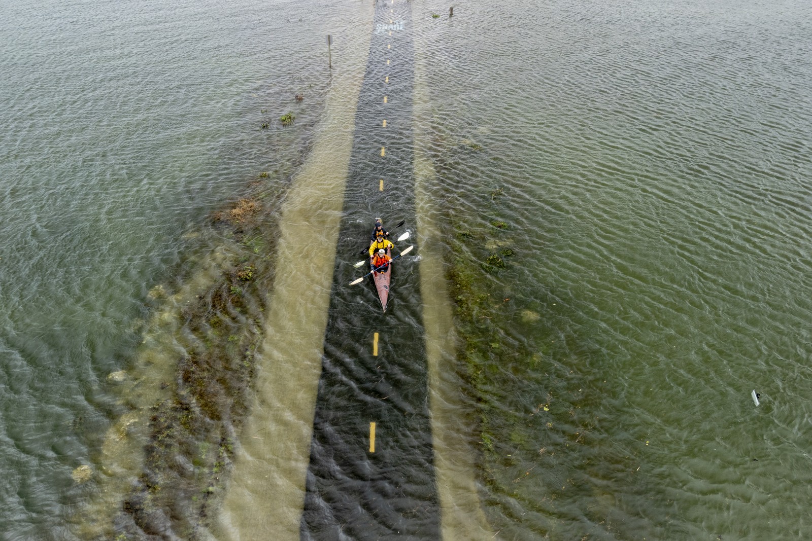 People in a three-person kayak paddle on shallow floodwater, showing a bike path just beneath the surface.