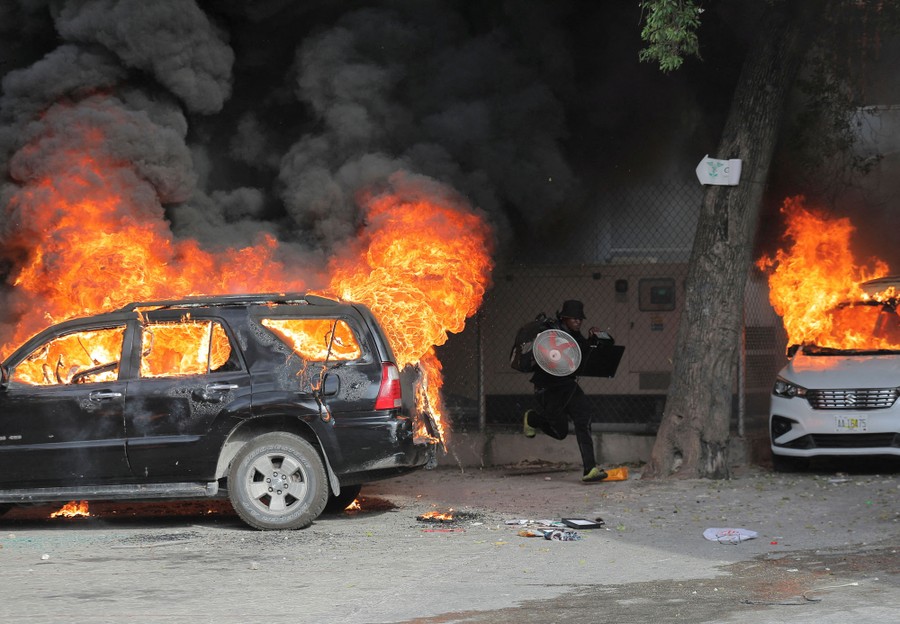 A person runs with looted goods near two burning cars.