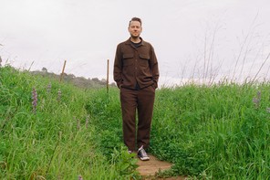Color photo of man in brown shirt and pants standing with hands in pockets on trail surrounded by green grass and wildflowers.