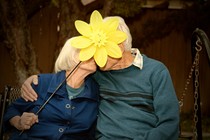 An older couple is kissing, and the woman is holding up a large yellow plastic lawn flower to hide their faces.