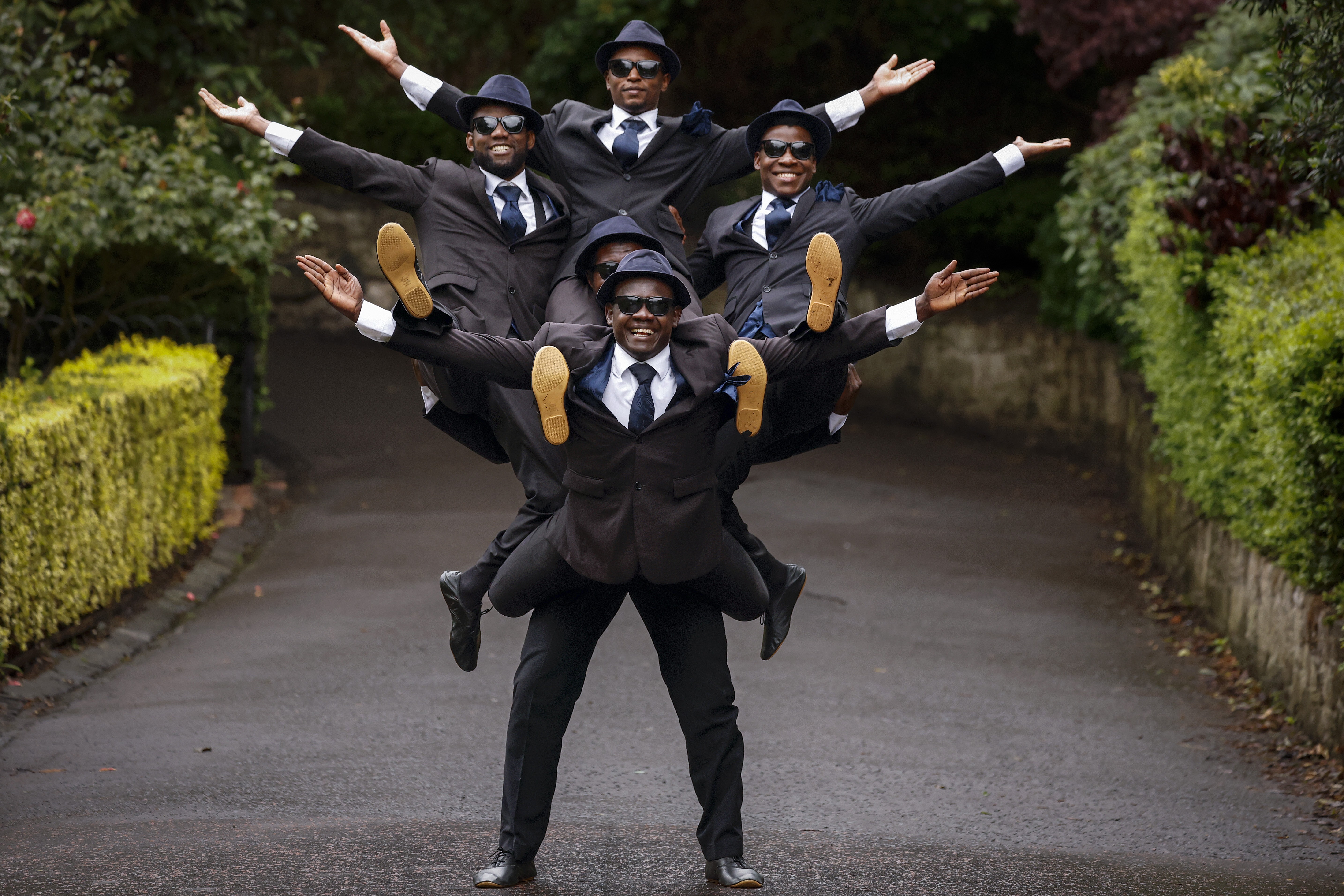 Four acrobats hang onto each other, supported by a fifth who stands in a street, as all raise their arms, posing.