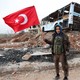 A Free Syrian Army fighter stands in front of a destroyed bus, holding a gun in one hand and a Turkish flag in the other.