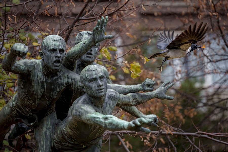 A bird flies past a statue of three men reaching forward, making it appear as if the bird is escaping the men.