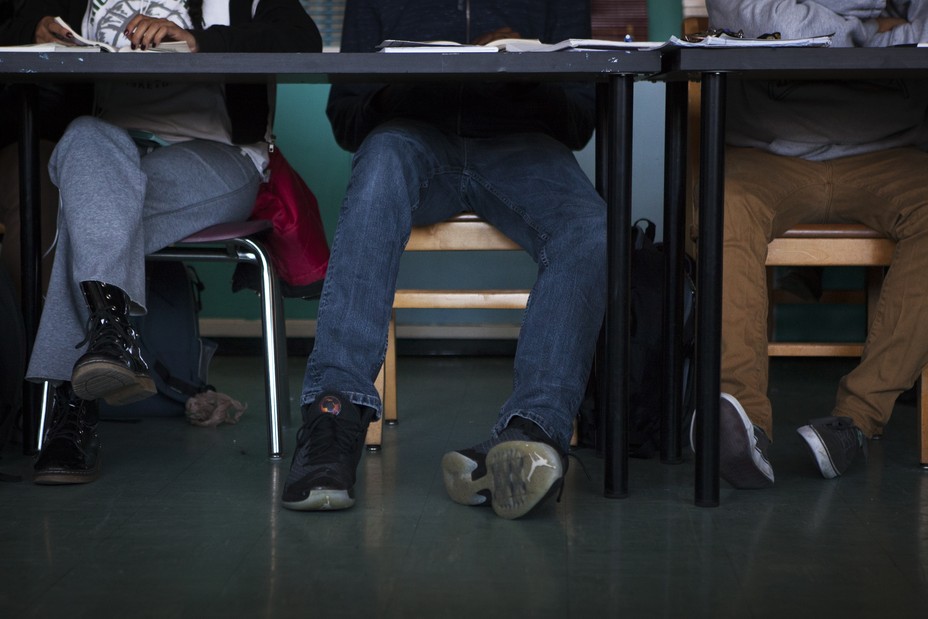 Students feet beneath their desks are shown. They wear jeans and sneakers. 
