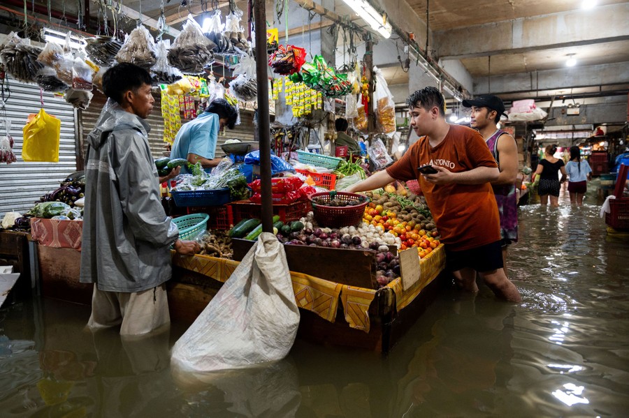 People shop in a flooded market, standing in knee-deep water.