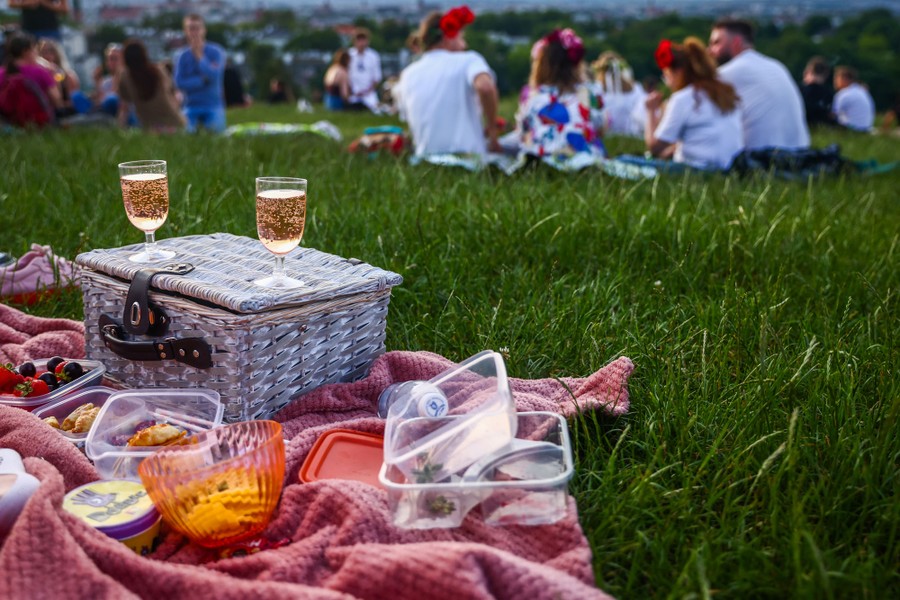 Food and drinks are seen on a blanket beside other picnickers nearby.