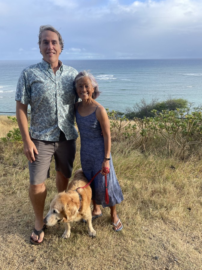 A tall middle-aged man and a short older woman pose with their arms around each other on a grassy beach with the ocean in the background. The woman is holding the leash of a golden retriever