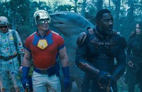 Five characters from "The Suicide Squad" stand together, facing forward. One wears a loose, gray bodysuit covered in colorful dots; another wears a silver helmet, a fitted red-and-blue T-shirt, and blue gloves; one is a human-shark hybrid; and two wear black.