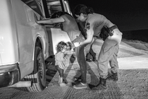 A black-and-white photo of a woman in a T-shirt and jeans leaning with both hands against a white van at night while a woman in uniform frisks her and a toddler stands underneath crying