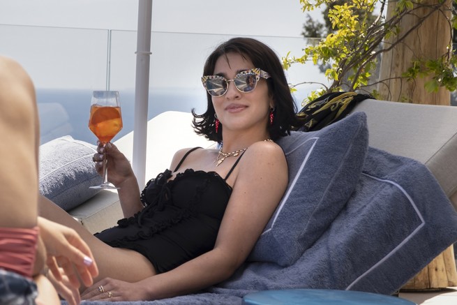 Simona Tabasco, as Lucia, wears sunglasses and reclines on a lounge chair with a drink in her hand.