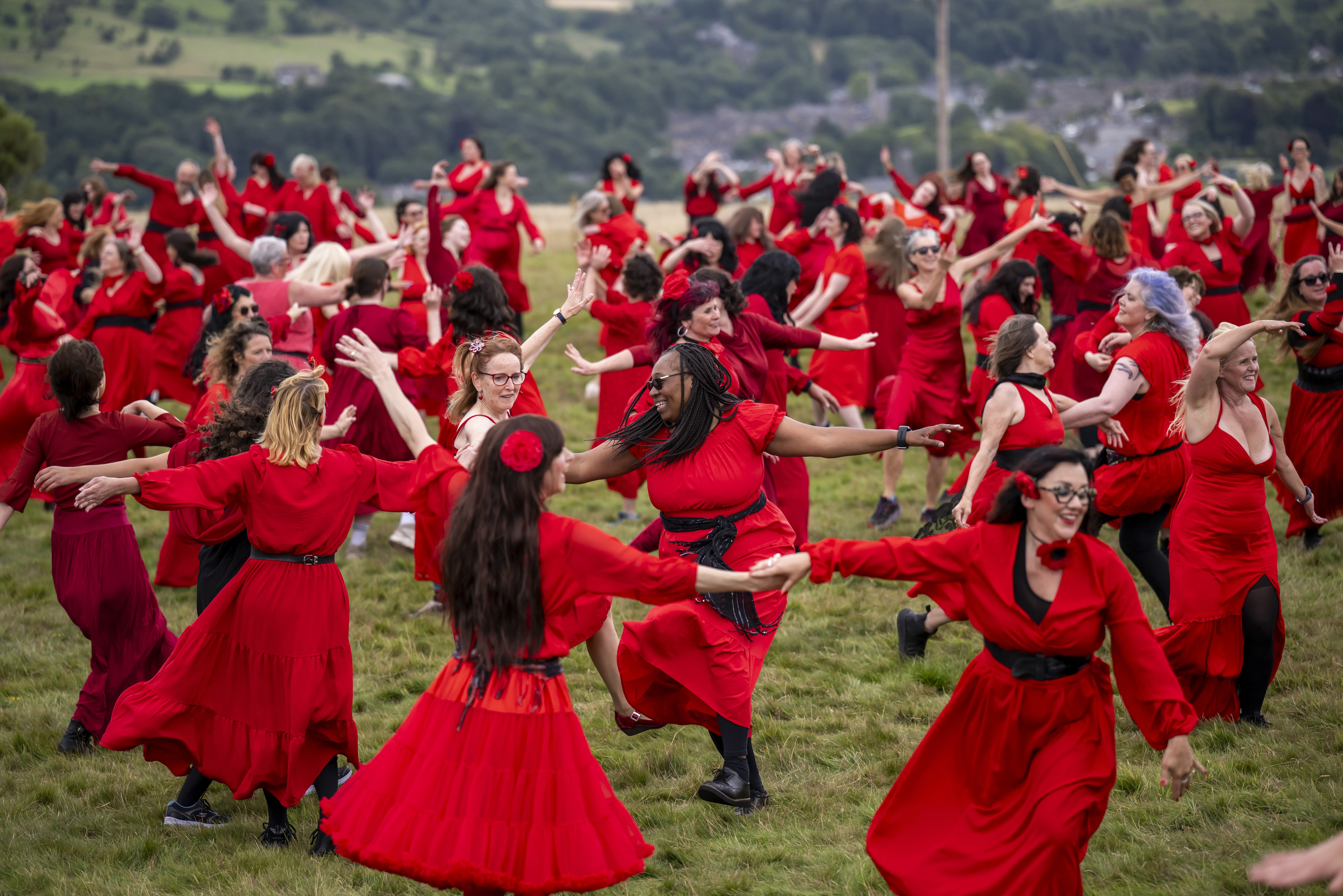 A large group of people, all wearing red dresses, dances in a field.