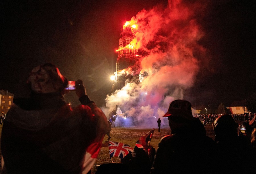 A crowd of people watch and take video as a very tall stack of wooden pallets goes up in flames.