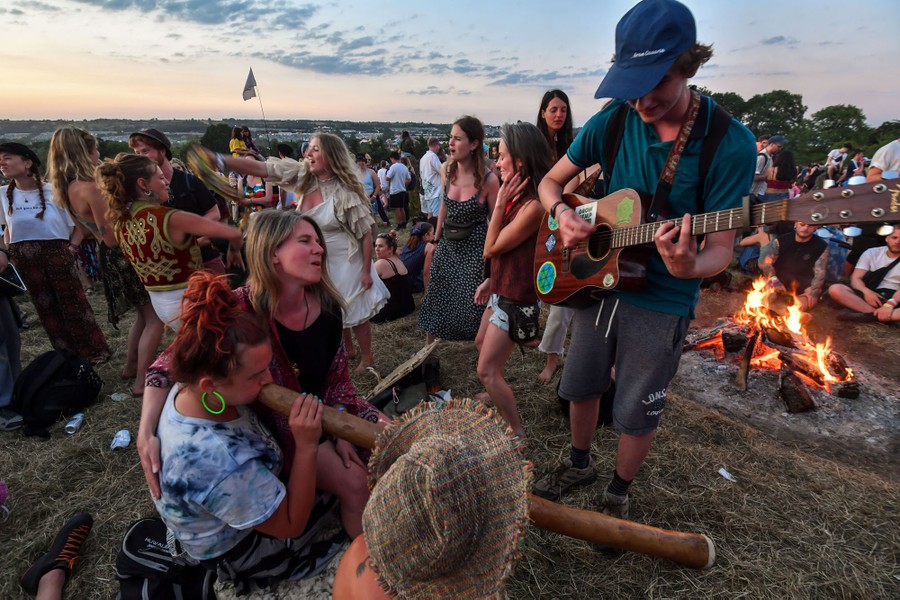 People gather in a field, playing music and dancing near a campfire.