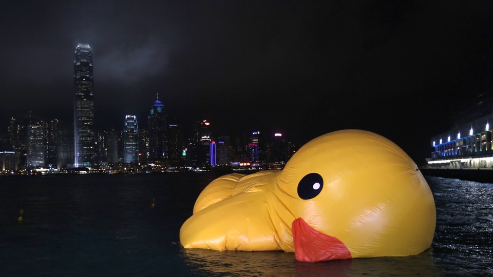 A partially deflated yellow duck float half-sunk in a river at night, with a city skyline in the background