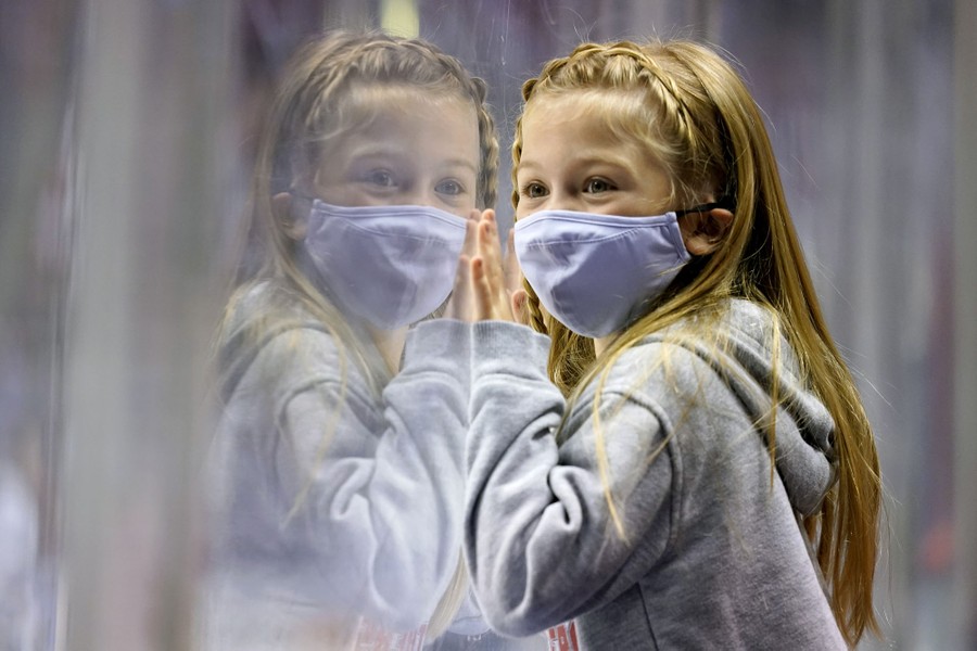 A girl wearing a face mask leans against a plexiglass barrier to watch hockey players.