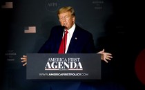 Donald Trump, wearing a dark suit, white shirt, and red tie, speaks at a lectern with an "America First Agenda Summit" banner across the front.