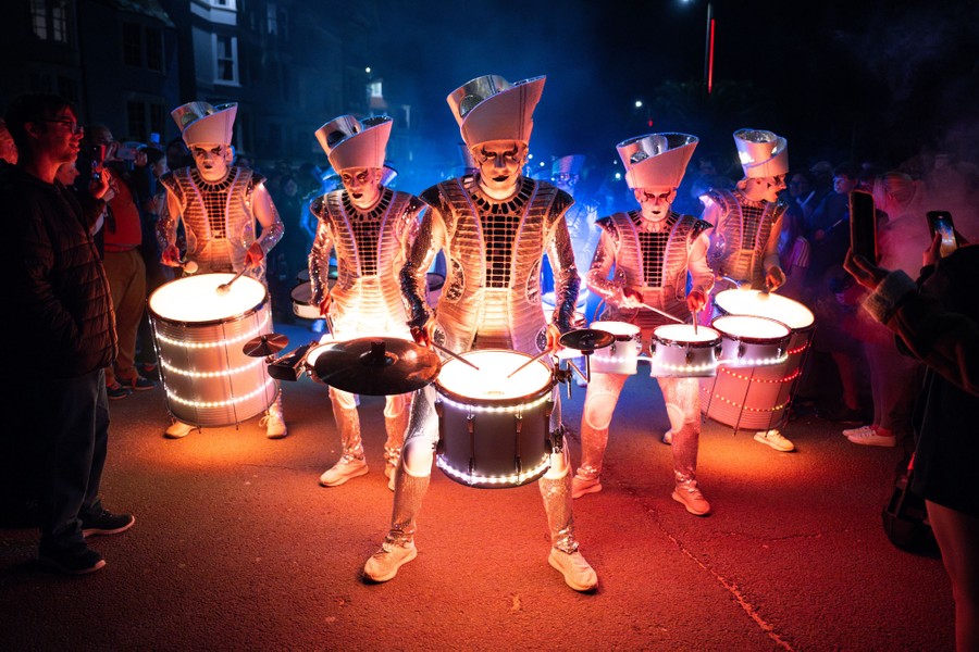 Five people in costume play illuminated drums in a street surrounded by a crowd.