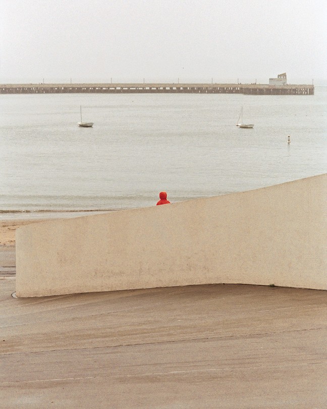 small figure in red hoodie sits by shore with 2 sailboats and pier in distance