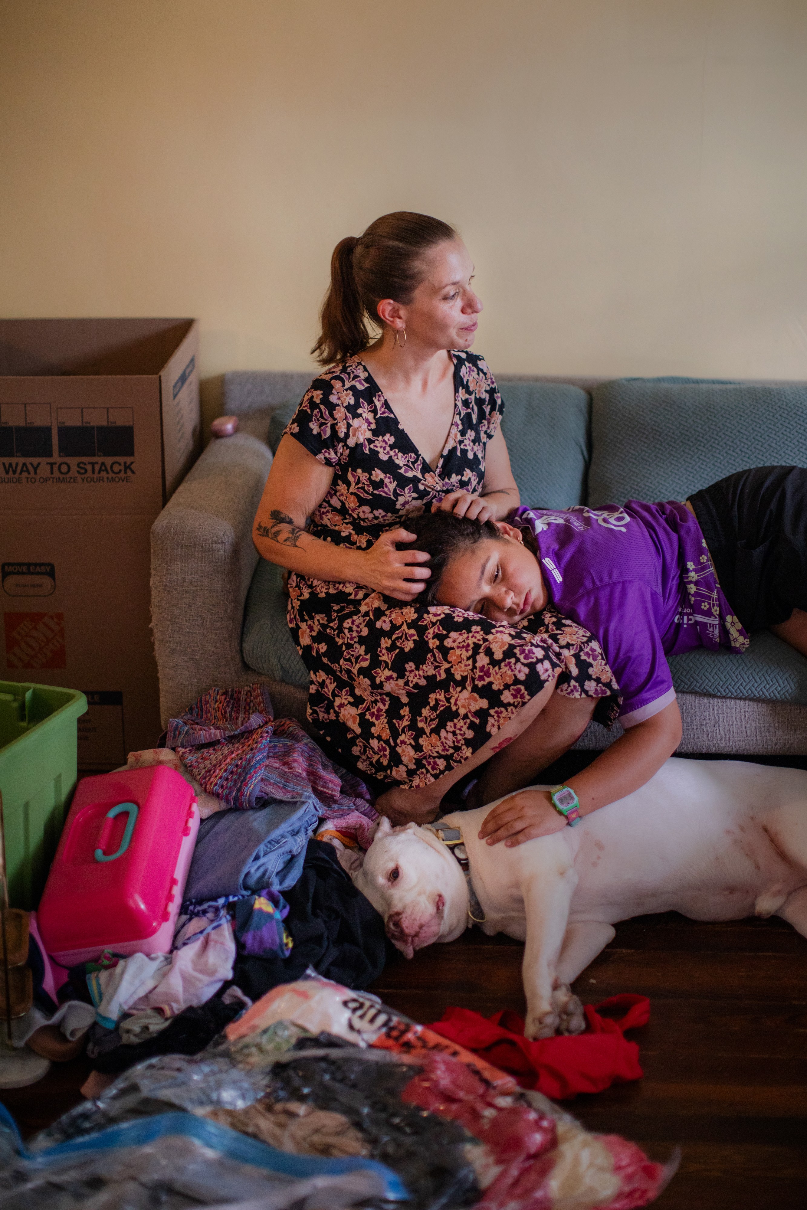 photo of woman surrounded by moving boxes sitting on sofa with daughter's head in her lap and arm petting a white dog lying on the floor