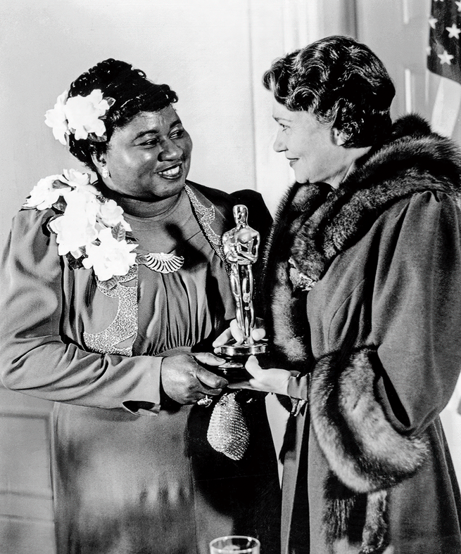 black-and-white photo of Hattie McDaniel in evening wear with flower in hair accepting Oscar statuette
