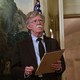 John Bolton holding a notepad and pen, standing in a formal room with a chandelier and U.S. flag in the background.