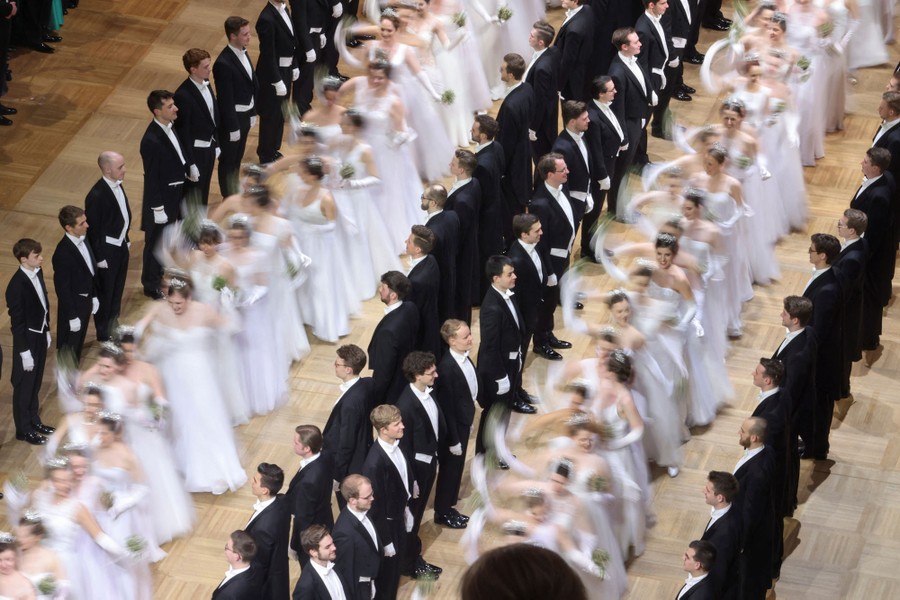 Several lines of men dressed in formal wear stand, watching two lines of women dancing in white gowns.