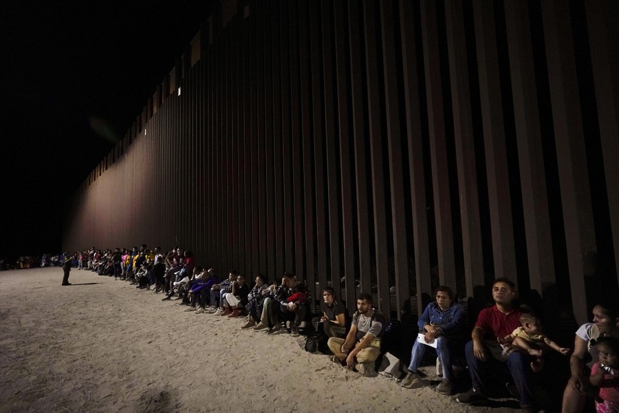 A long line of people sit against a tall steel border wall.