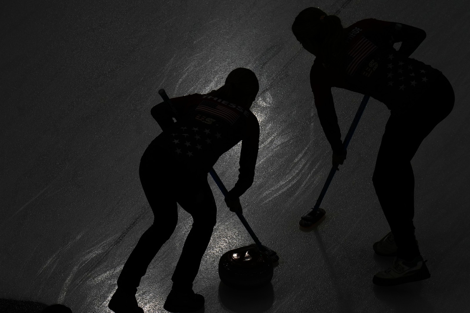 Two athletes compete during a curling match, seen in silhouette on ice.