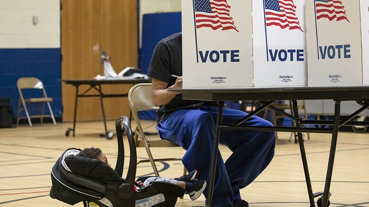 A child in a carrier sits next to their father as he votes. The father's face is covered by a partition that reads "VOTE" and is decorated with an American flag.