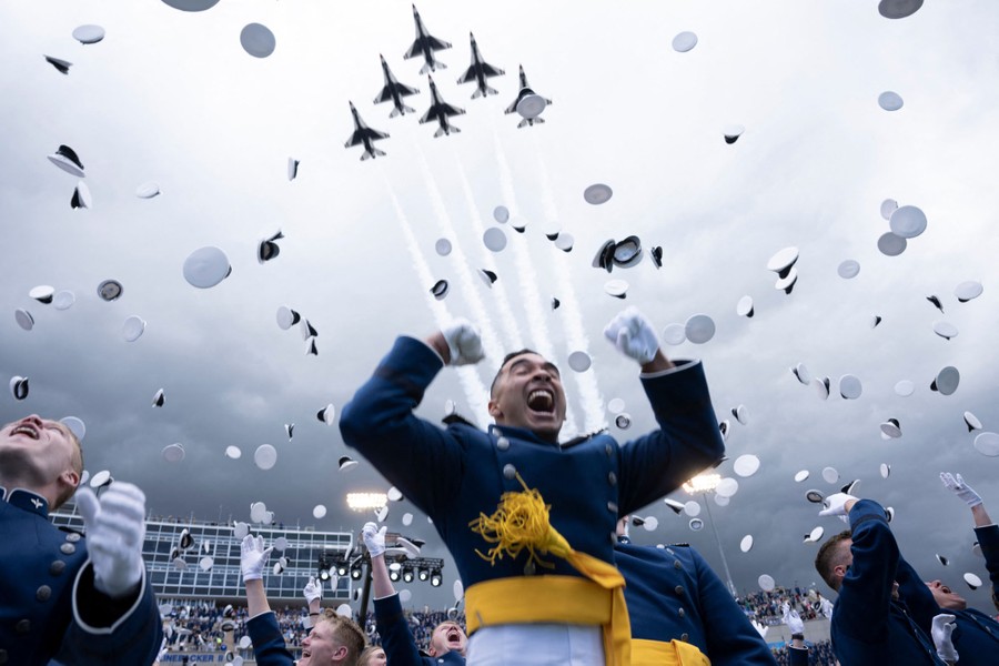 Graduates in dress uniforms cheer and throw their hats in the air as six jets fly above in formation.
