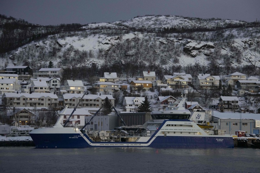 A view from the water of a large fishing vessel docked near a residential neighborhood on a snowy hillside.