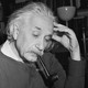 A black and white photograph of Albert Einstein at a desk, pipe in mouth, left hand against his temple