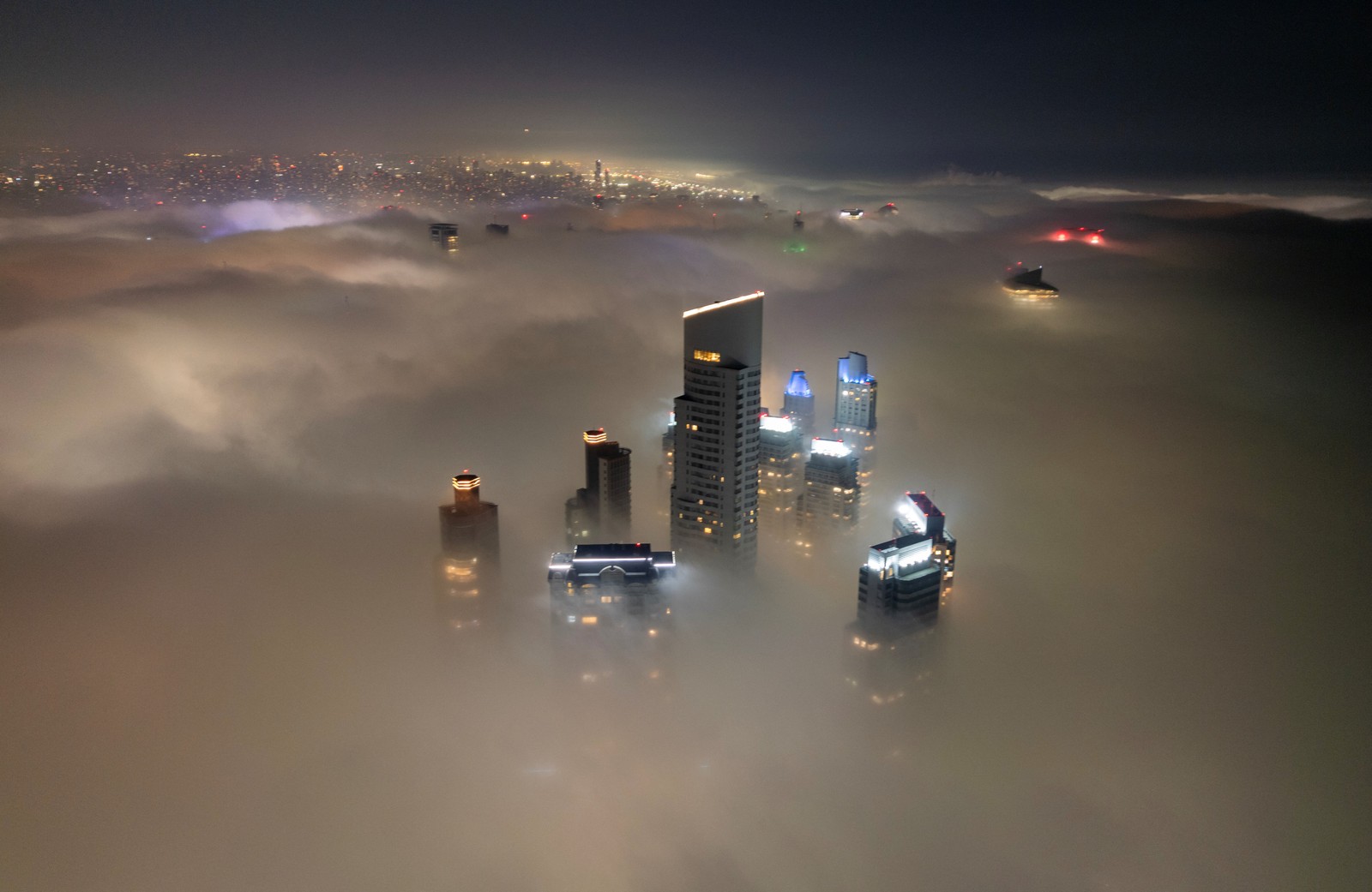 The tops of several skyscrapers poke above a bank of low clouds at night.
