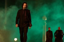 Keanu Reeves, as John Wick, walks toward the camera, green smoke billowing around him and a stoic expression on his face.