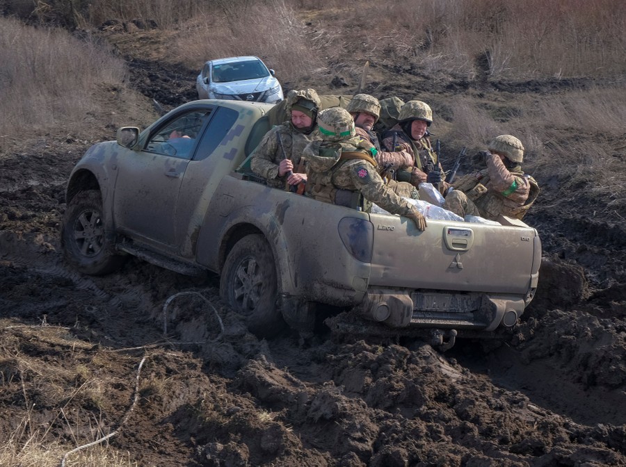 Five soldiers ride in the back of a small pickup truck on a very muddy road.