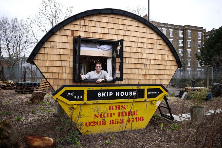 A person leans out the window of a tiny house built into a dumpster.
