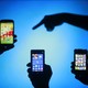 Three silhouettes of hands holding smartphones with silhouette of finger pointing at them on blue background