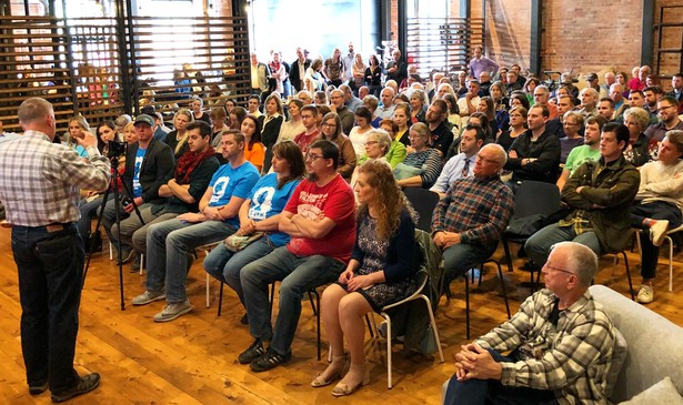 A man at the front of the room speaks to a crowded room, with rows of seated people and more standing in the back to listen