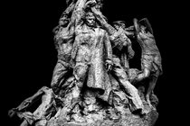 A black-and-white photo of a statue of a group of people standing, hands stretching toward the sky