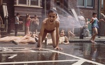 Children play in a city in front of rowhouses, in their swimwear, as a fire hydrant sprays water. One boy, front and center, seems excited and grins directly at the camera.