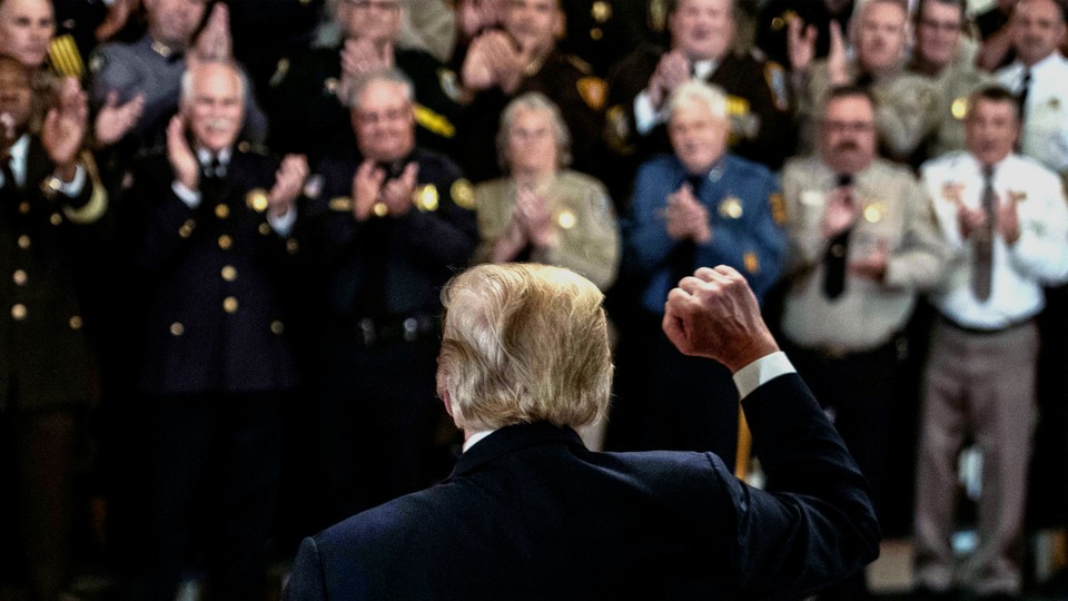 A photograph of Donald Trump from behind, looking out over a crowd of law-enforcement officials, with his fist raised